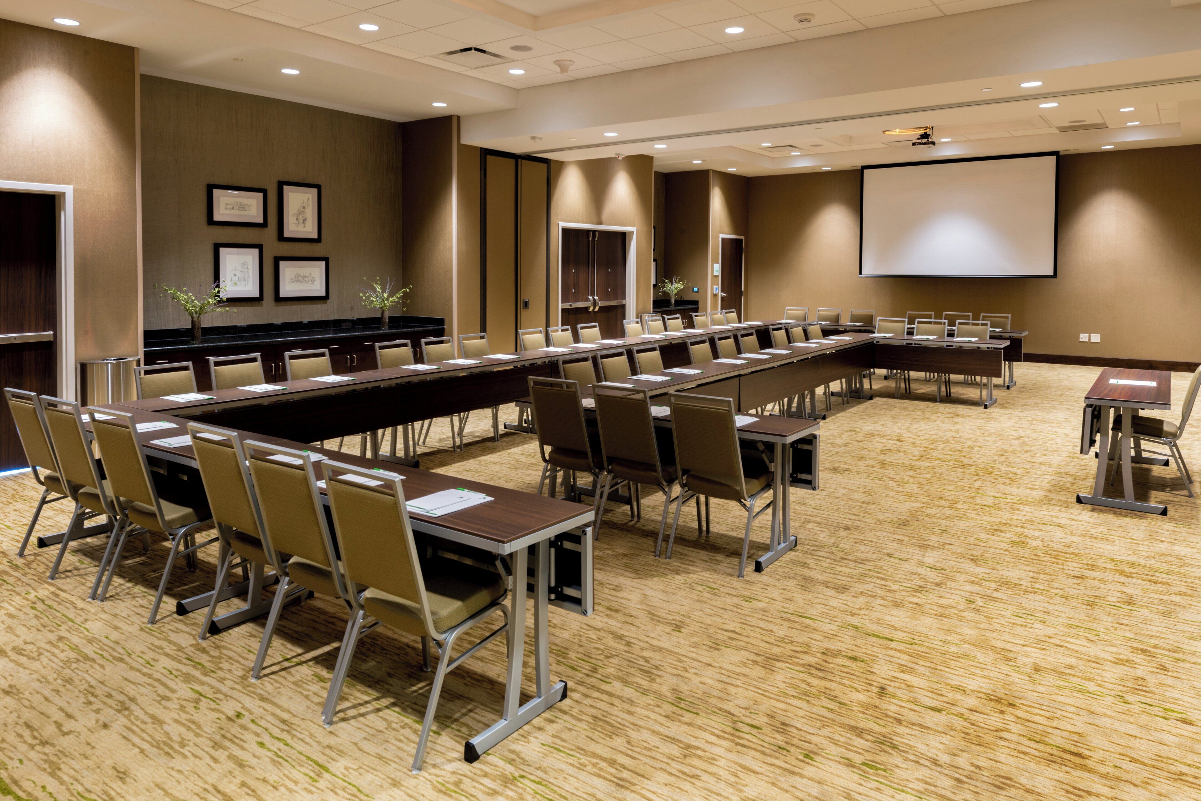 A conference room with a long table, chairs, a projector screen, and framed artwork on the walls.