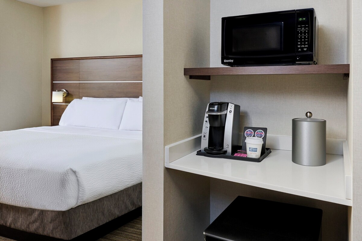 A hotel room with a bed, a minibar, a microwave, a coffee maker, and a trash bin.