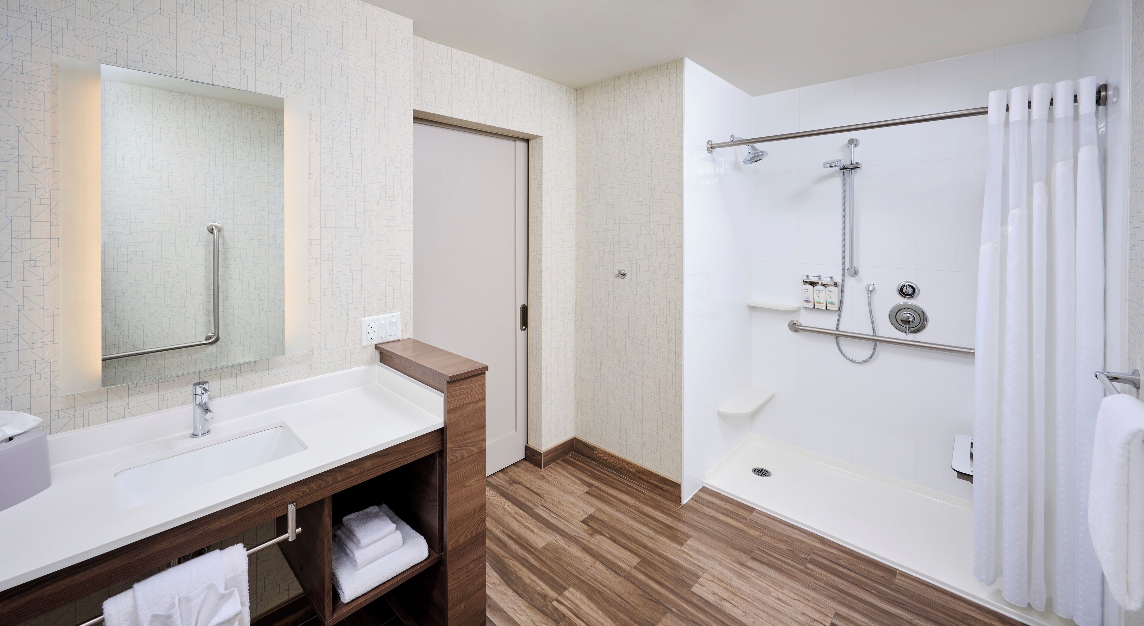 A modern bathroom with a wooden vanity, a large mirror, a walk-in shower with grab bars, and a white tiled floor.