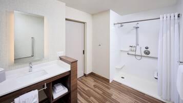 A modern bathroom with a wooden vanity, a large mirror, a walk-in shower with grab bars, and a white tiled floor.
