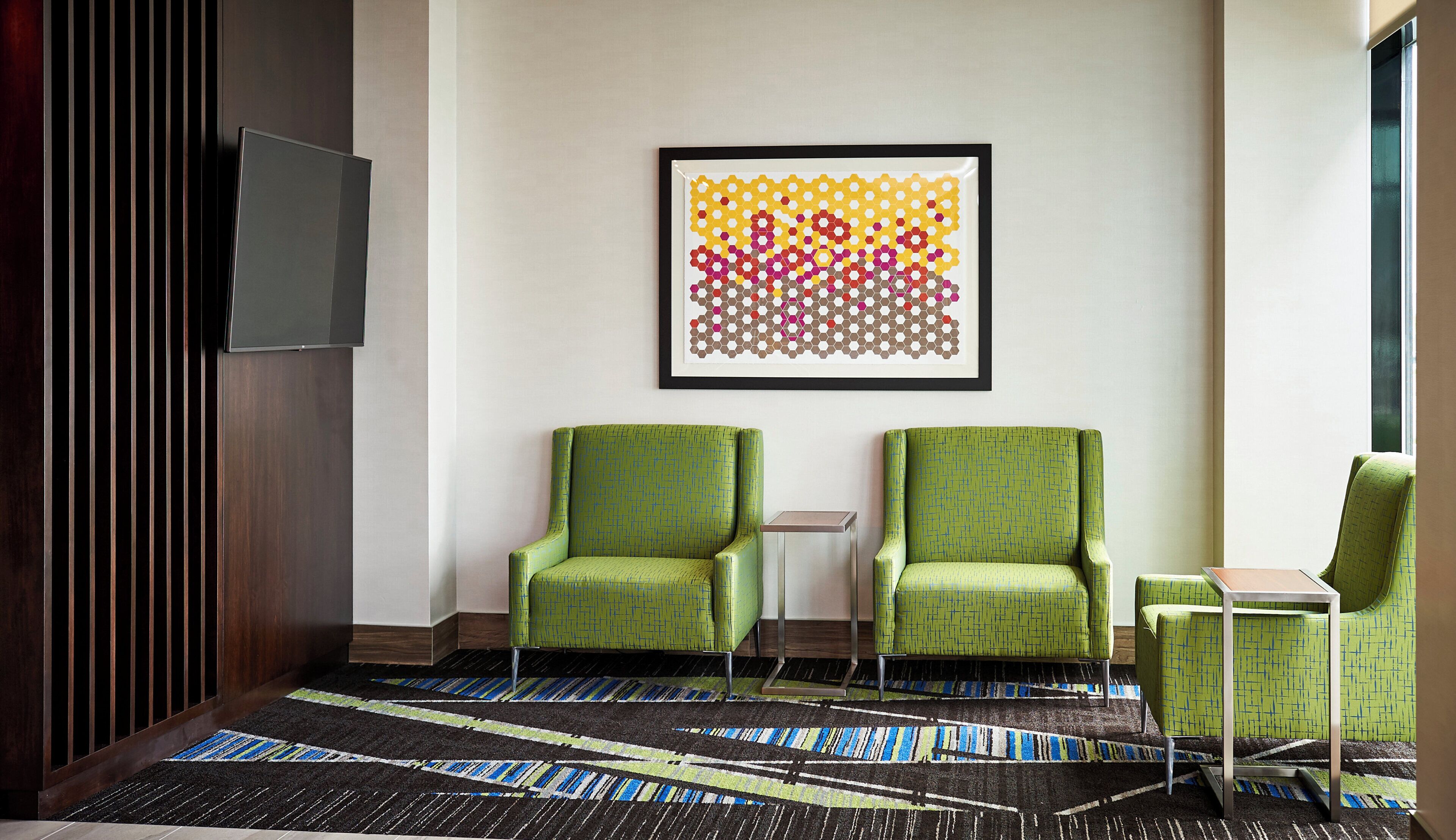A hotel lobby with two green armchairs, a small table, a framed abstract art piece, a flat-screen TV, and a patterned carpet.