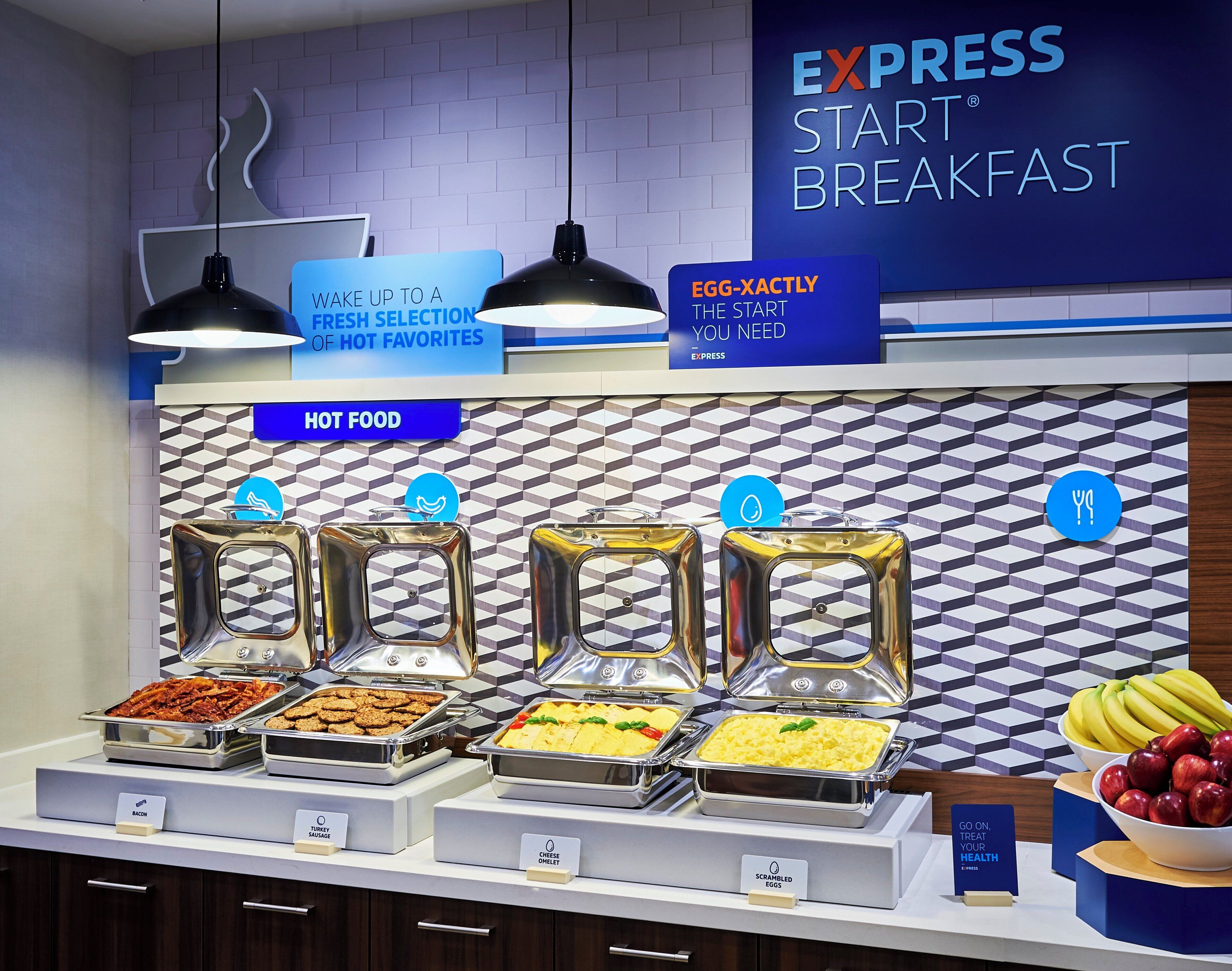 Een ontbijtbuffet met aangeduide stations voor eten, waaronder spek, kalkoenworstjes, kaasomelet, roerei en een schaal bananen en appels.