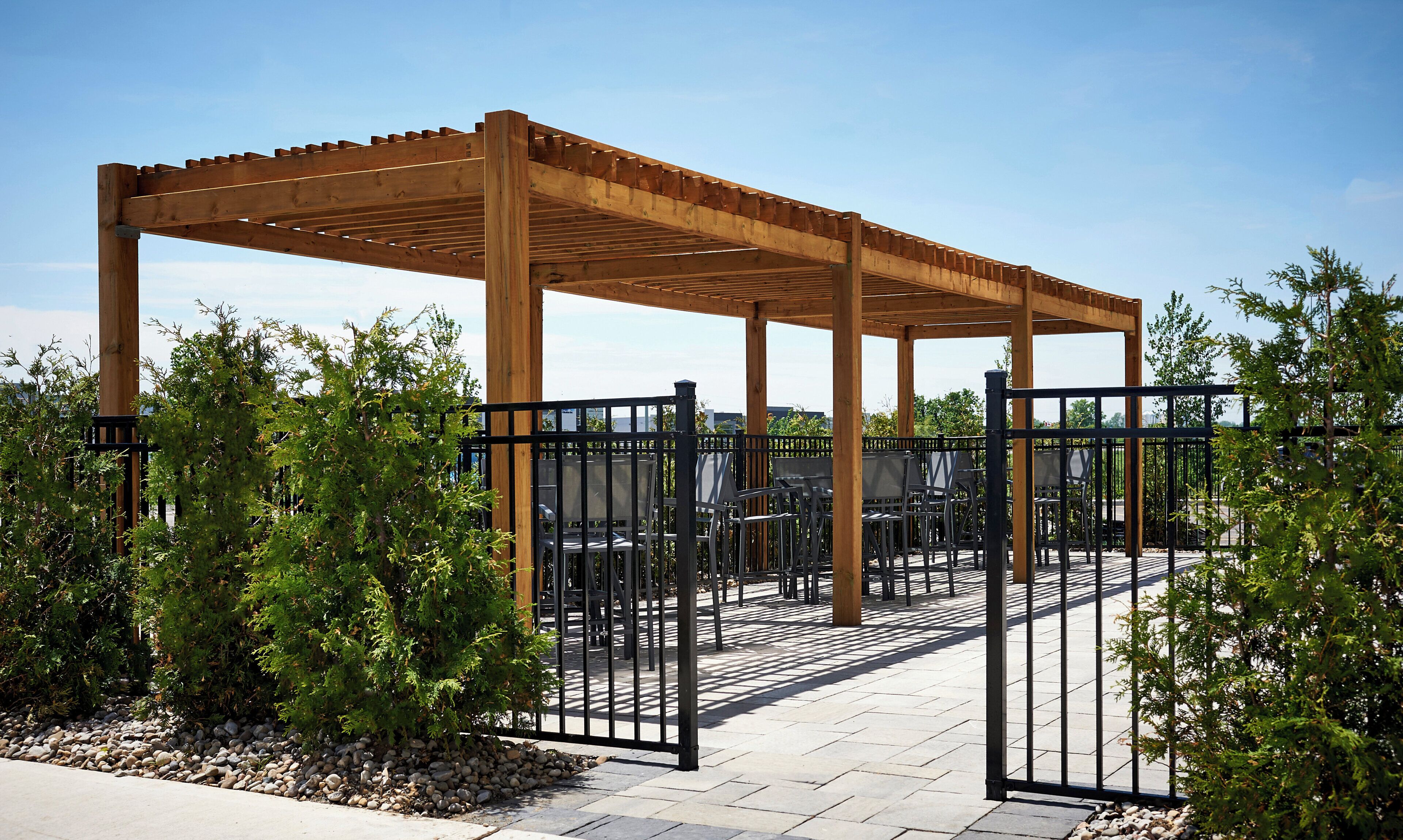 En pergola i træ med siddepladser under, bestående af stole og borde, omgivet af et hegn og grøn bevoksning.