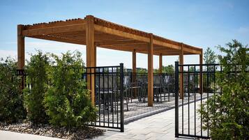 En pergola i træ med siddepladser under, bestående af stole og borde, omgivet af et hegn og grøn bevoksning.
