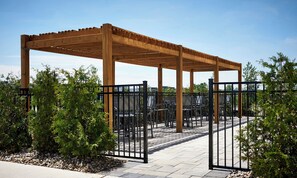 A wooden pergola structure with metal chairs and tables underneath, surrounded by a metal fence and greenery.