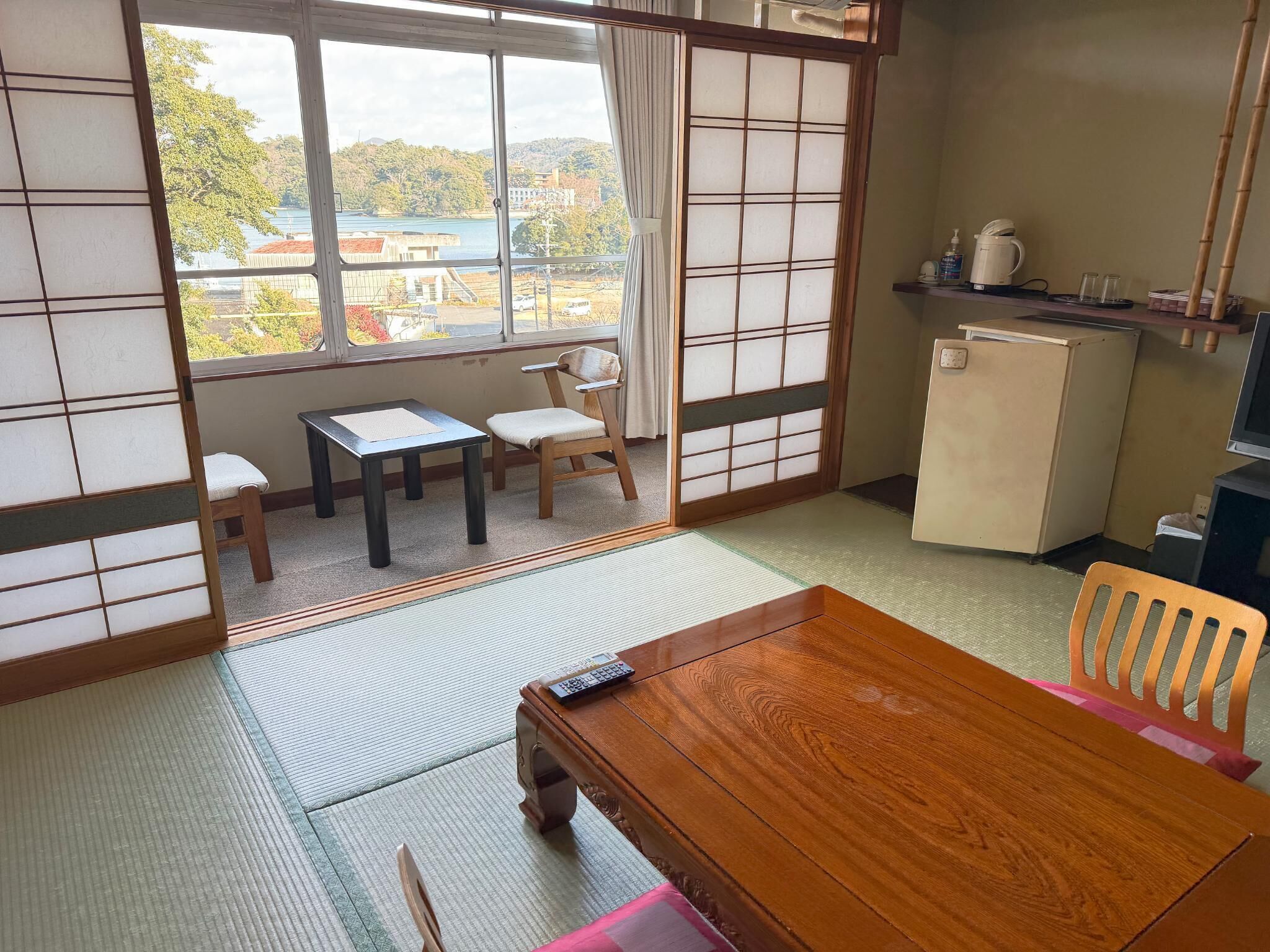 Una habitación japonesa tradicional con suelo de tatami, una mesa de madera, una silla, una mesita auxiliar, una puerta corredera y vistas a un lago y edificios.
