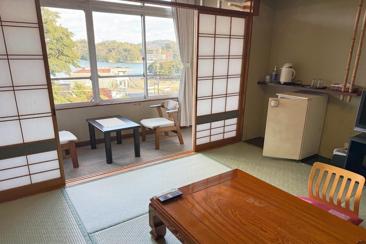 Une pièce de style japonais traditionnel, avec un sol recouvert de tatamis, une table en bois, une chaise, une petite table d’appoint, une porte coulissante et une vue sur un lac et des bâtiments.