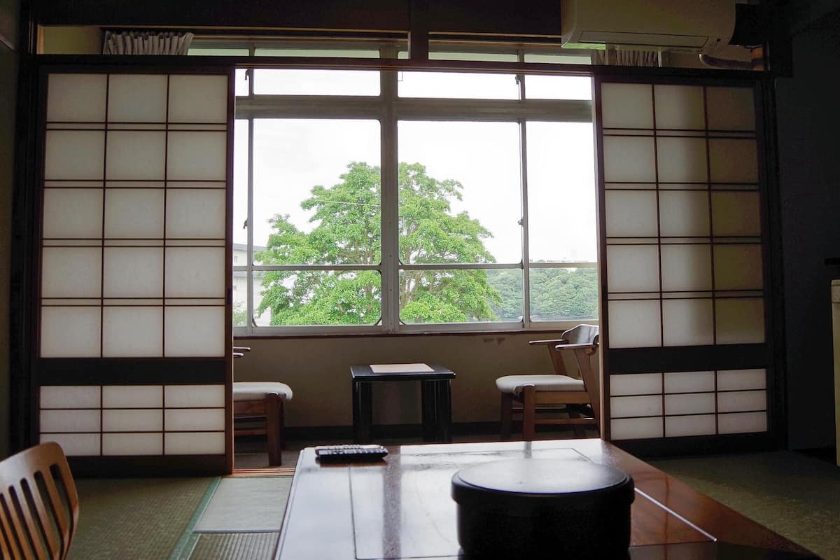 Une pièce de style japonais traditionnel, avec un sol recouvert de tatamis, une petite table basse, deux chaises et une fenêtre donnant sur la forêt.