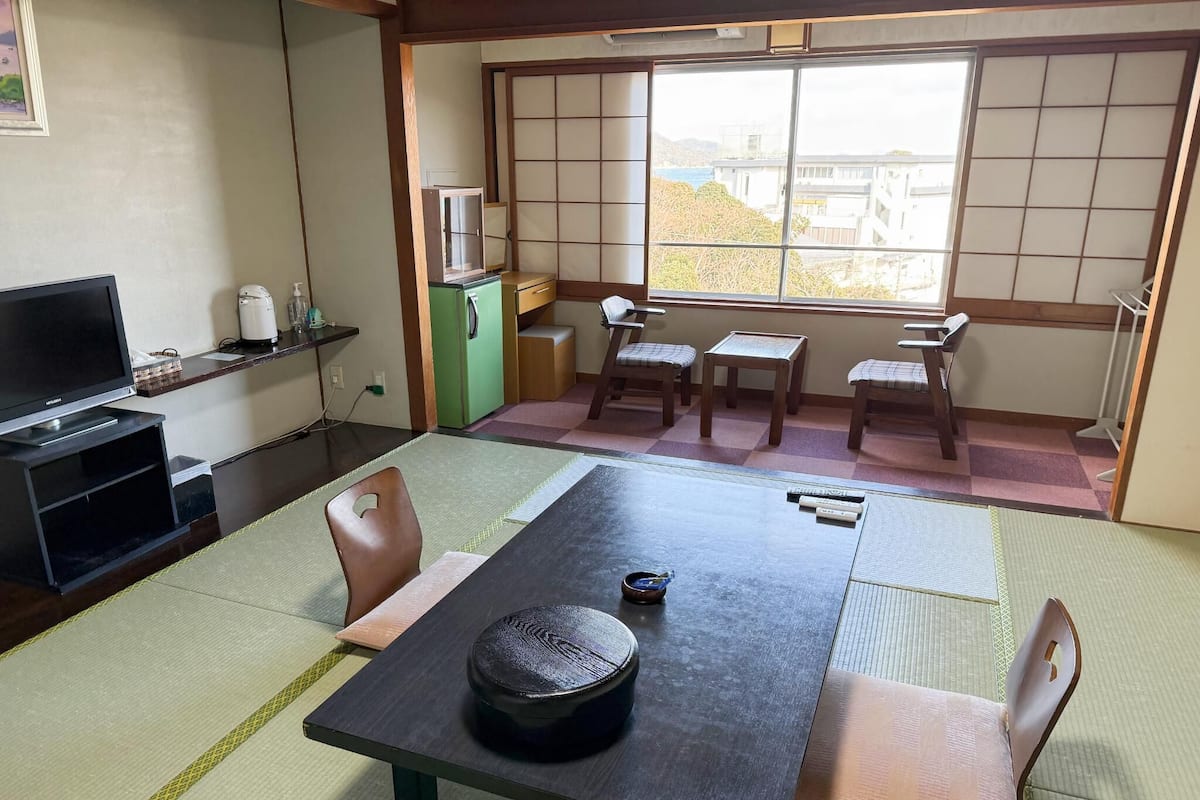 Une pièce de style japonais traditionnel, avec un sol recouvert de tatamis, une petite table sur laquelle repose un plateau noir, et un téléviseur posé sur une étagère.