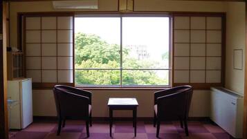 A room with tatami flooring, a low table, two chairs, and a large window with a view of greenery.