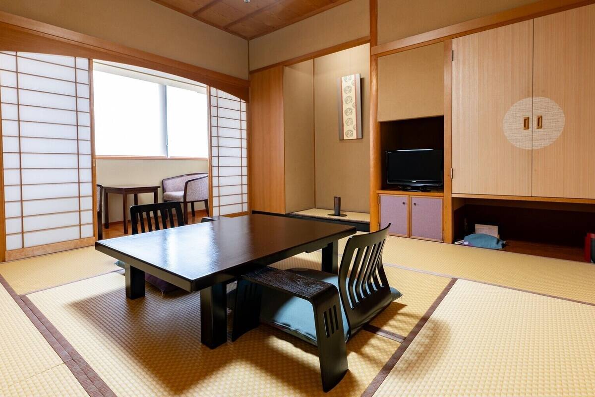 A traditional Japanese room with tatami flooring, a low table, chairs, a sliding door, and a television cabinet.