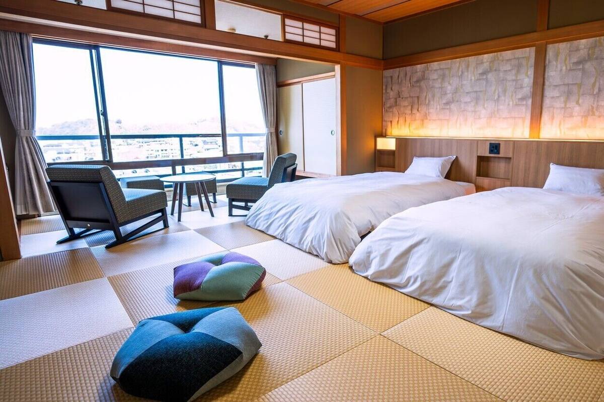 A hotel room with two beds, tatami mats, a view through large windows, and a traditional Japanese-style wall.