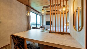 A modern hotel room with a wooden dining table, two chairs, a bed, a TV, and a large window with a view.