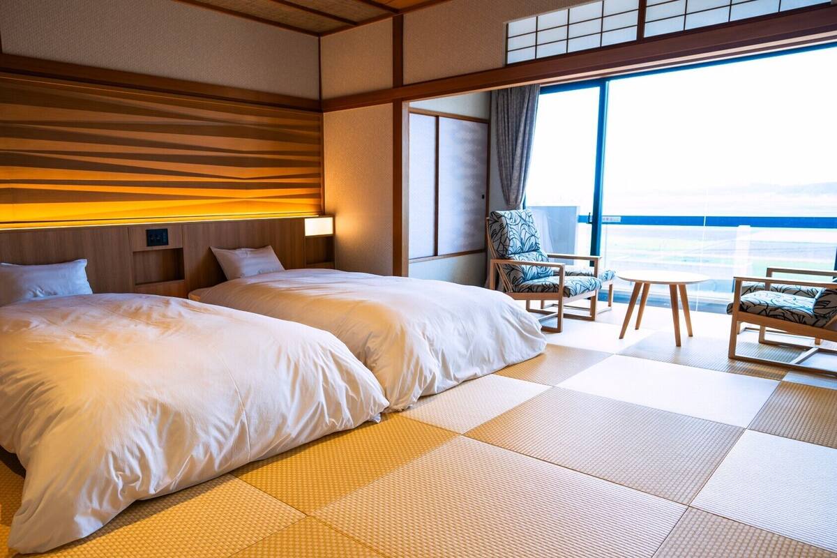 A hotel room with two beds, a chair, a small table, and a large window offering a view.