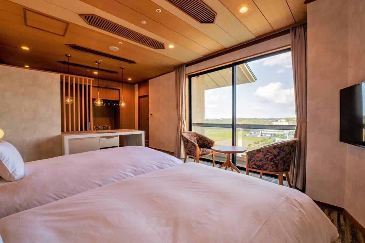 A hotel room with a large bed, a desk, two chairs, a TV, and a large window with a view.