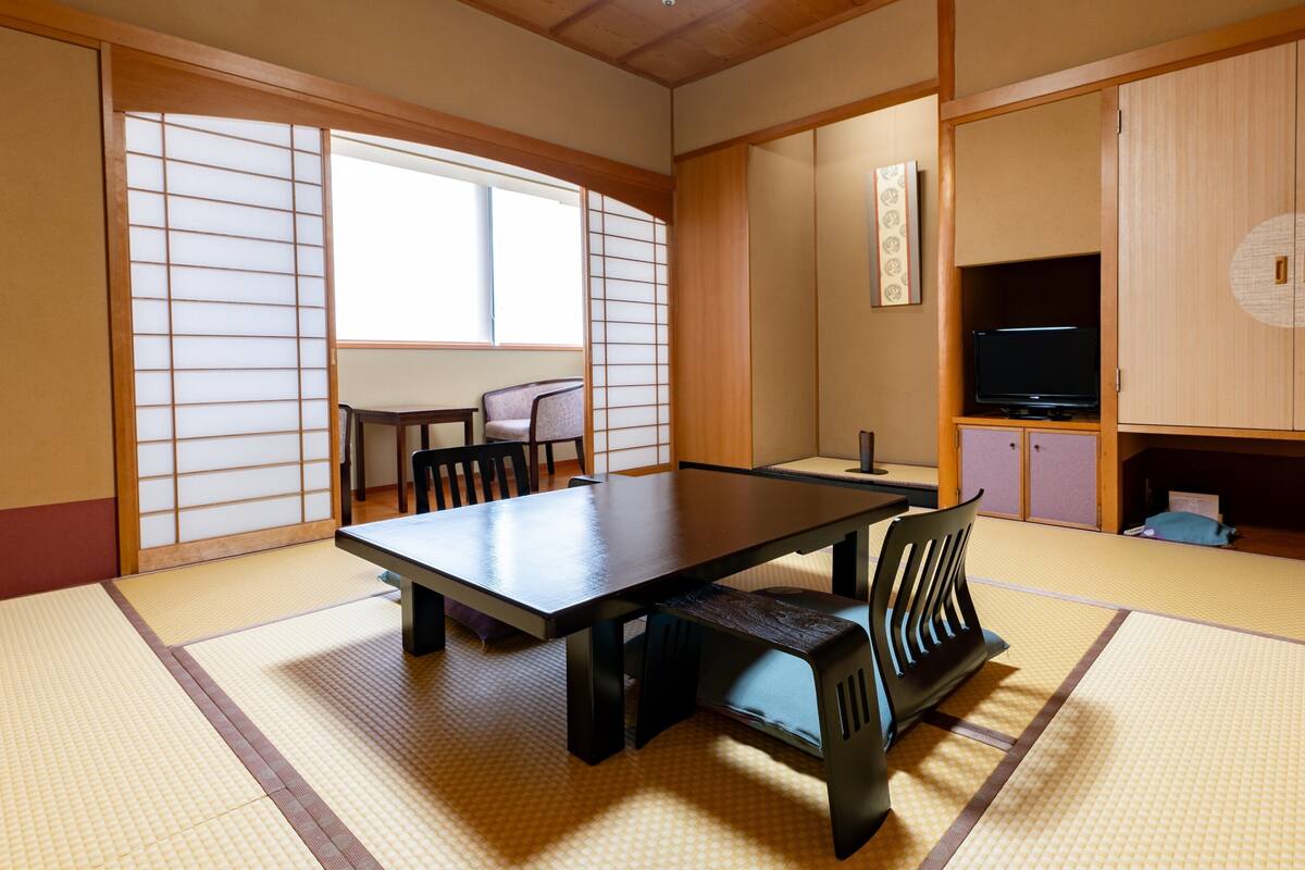 A traditional Japanese room with tatami flooring, a low table, chairs, a television, and sliding doors.