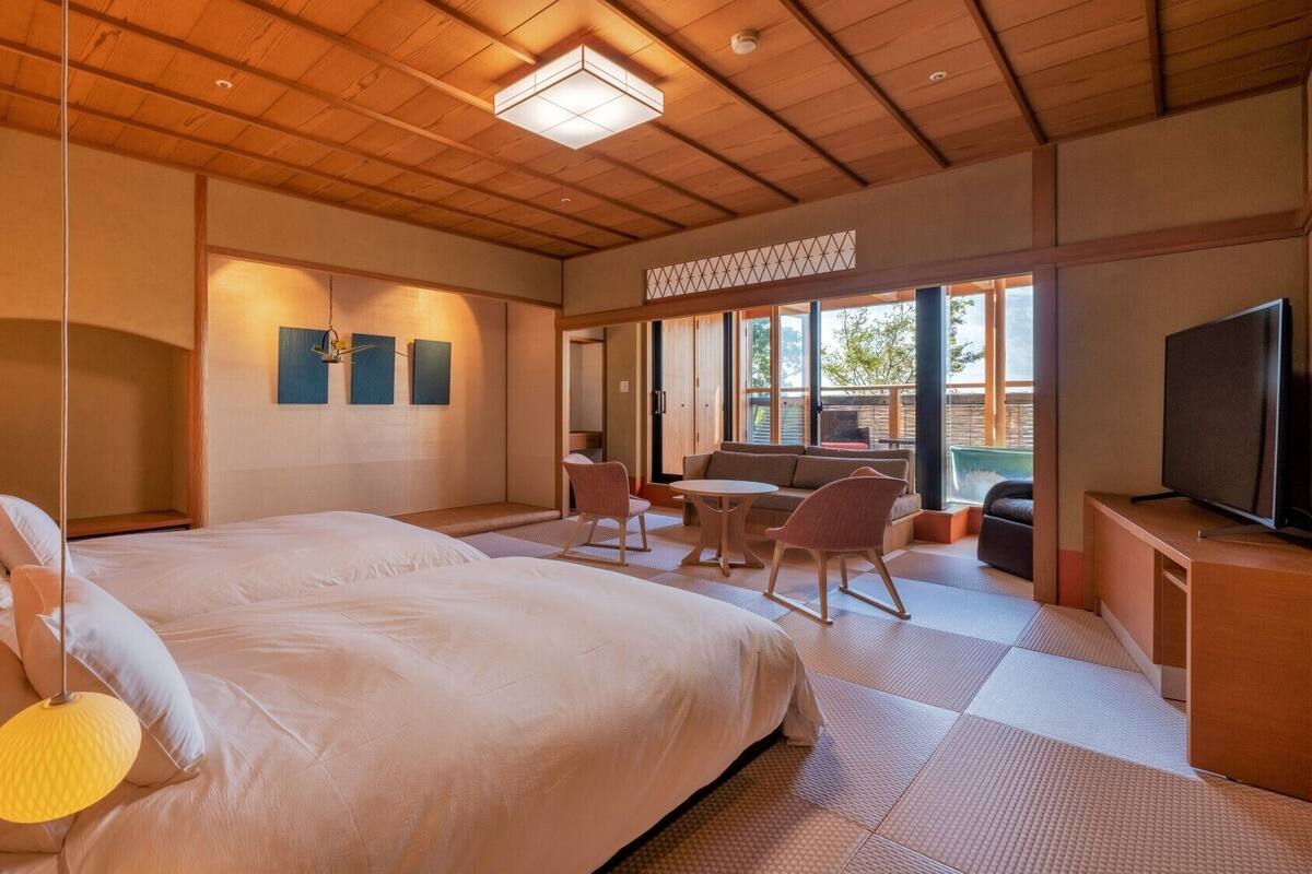 A hotel room with a large bed, a TV, a dining area, and a view of the outdoors.