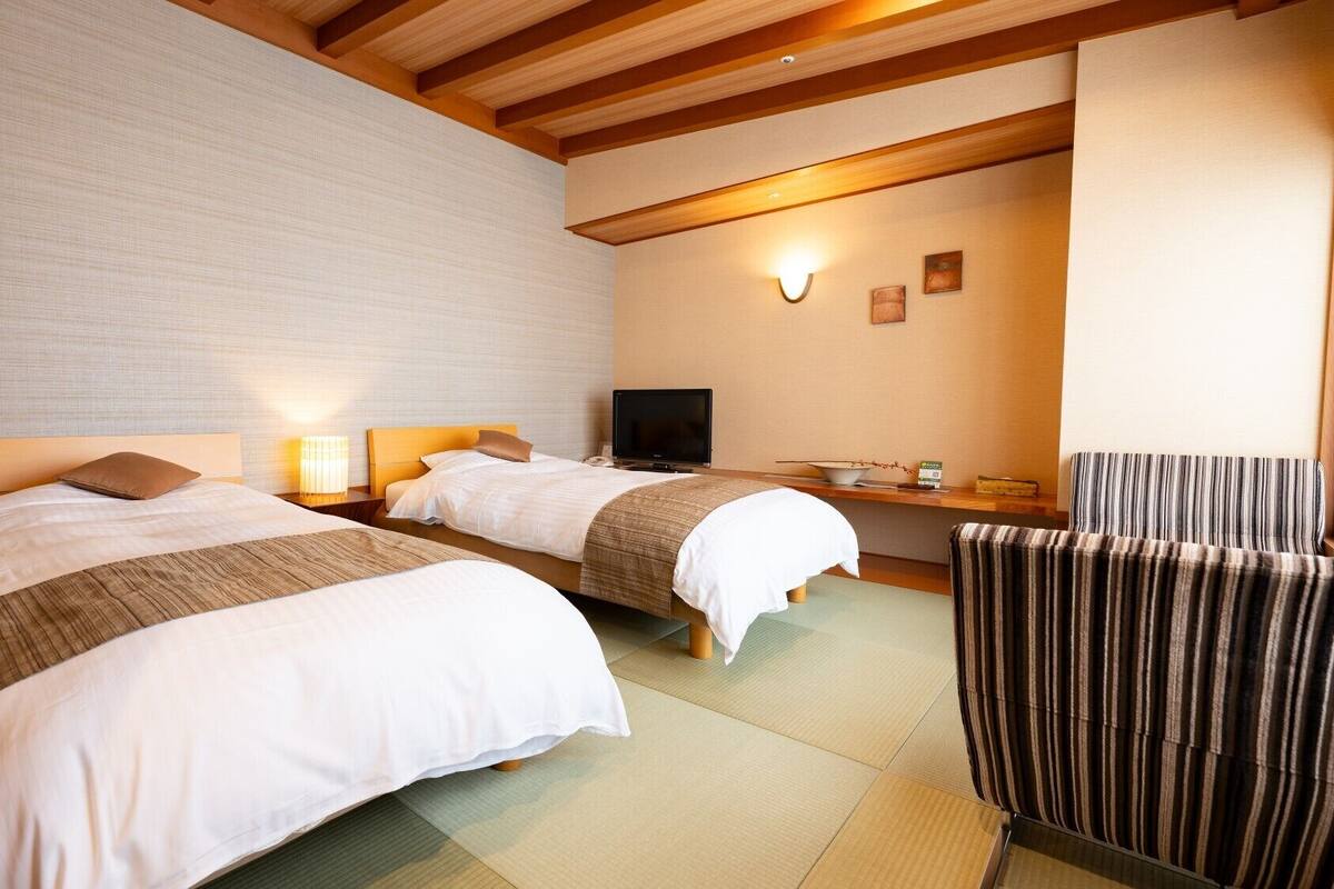 A hotel room with two beds, a TV, a chair, and a small table with a bowl and a book.