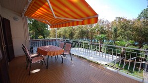 A balcony with a table and chairs, a striped awning, and a view of trees and a road. - Holiday apartment with balcony (Njivice)