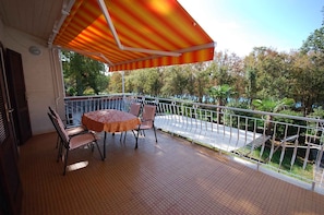 Un balcone con tavolo e sedie, un tendaggio a righe e vista su alberi e una strada.
