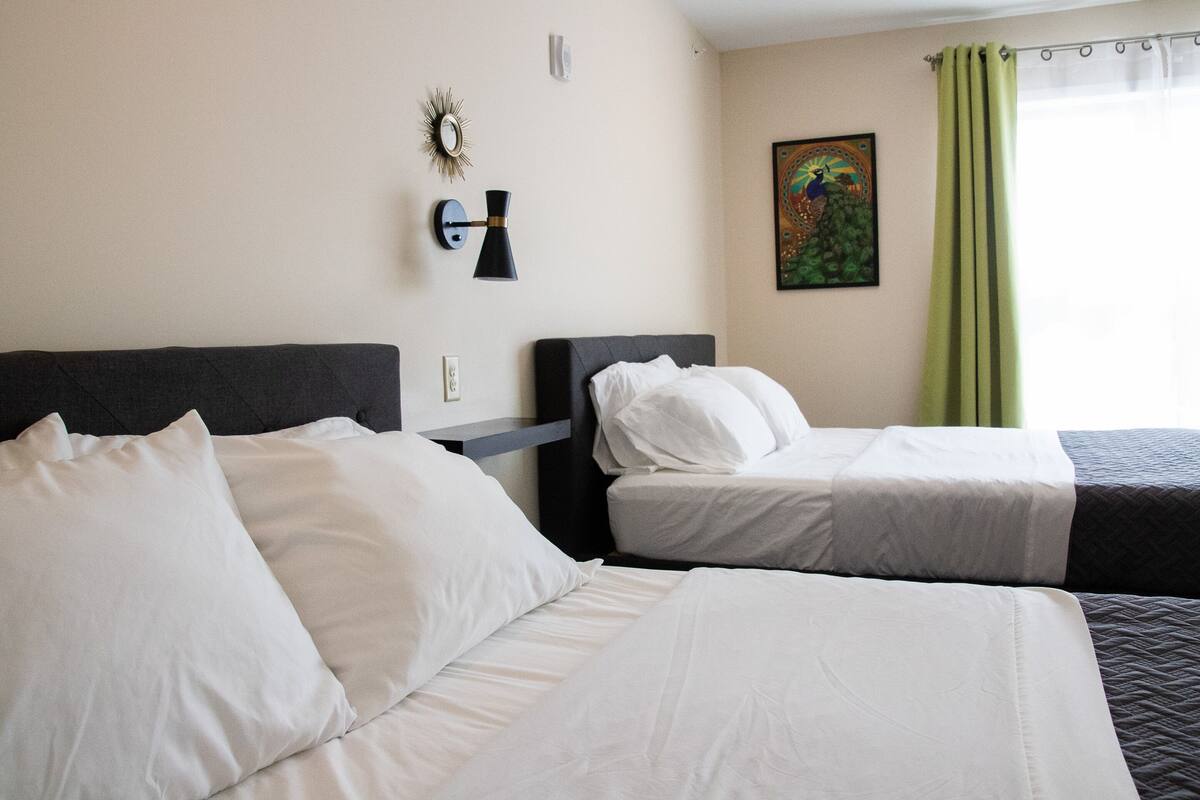 A hotel room with two beds, a wall-mounted lamp, a painting, and a window with curtains.