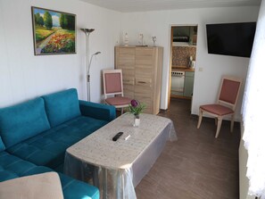 Un salon avec un canapé bleu, une petite table sur laquelle est posé un vase, un meuble en bois et, au fond, un coin cuisine.
