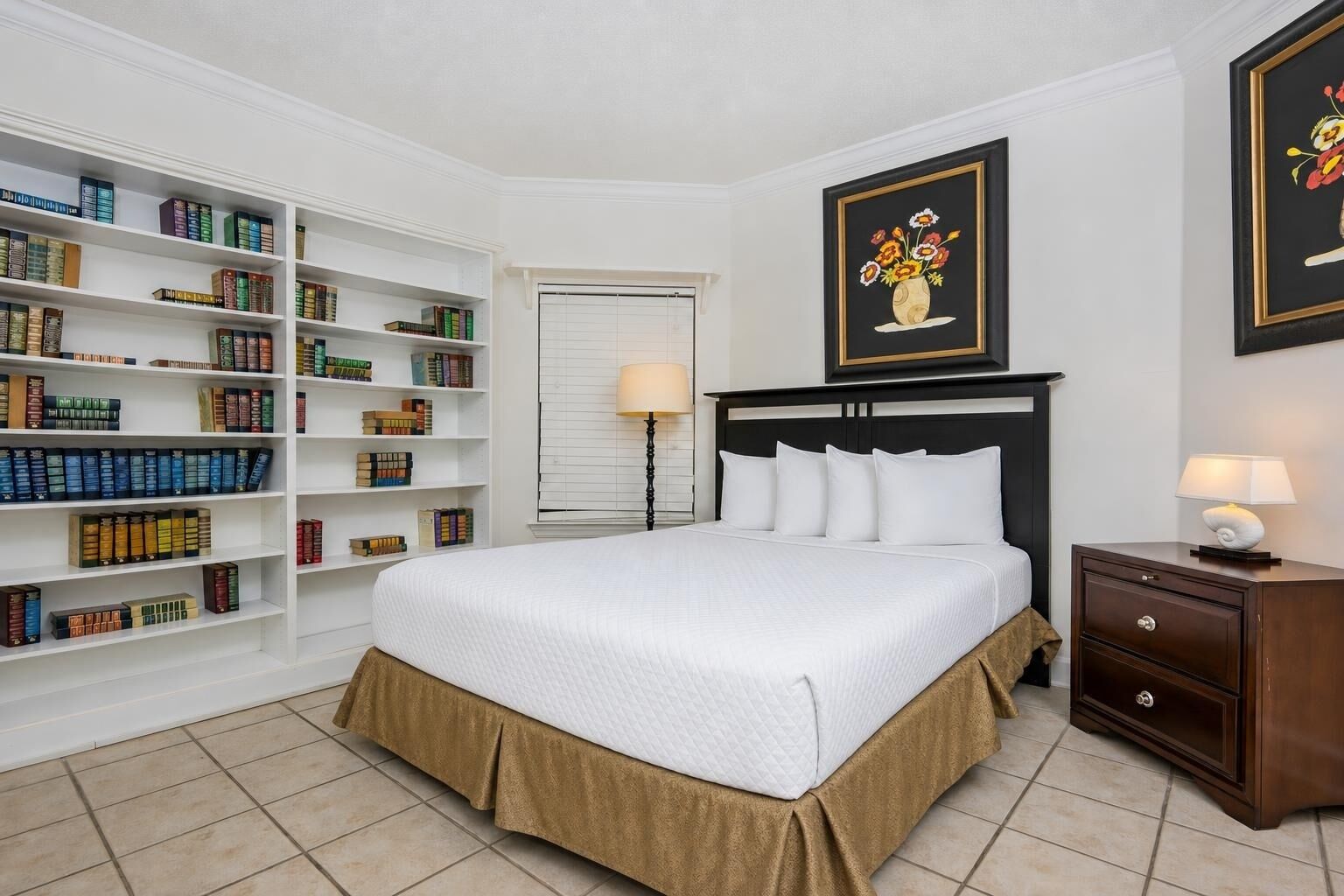 A hotel room with a large bookshelf, a bed with white bedding, a nightstand with a lamp, and a framed floral artwork on the wall.