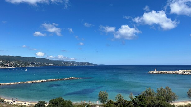 A coastal view with clear skies, a rocky breakwater, and a distant shoreline with buildings.