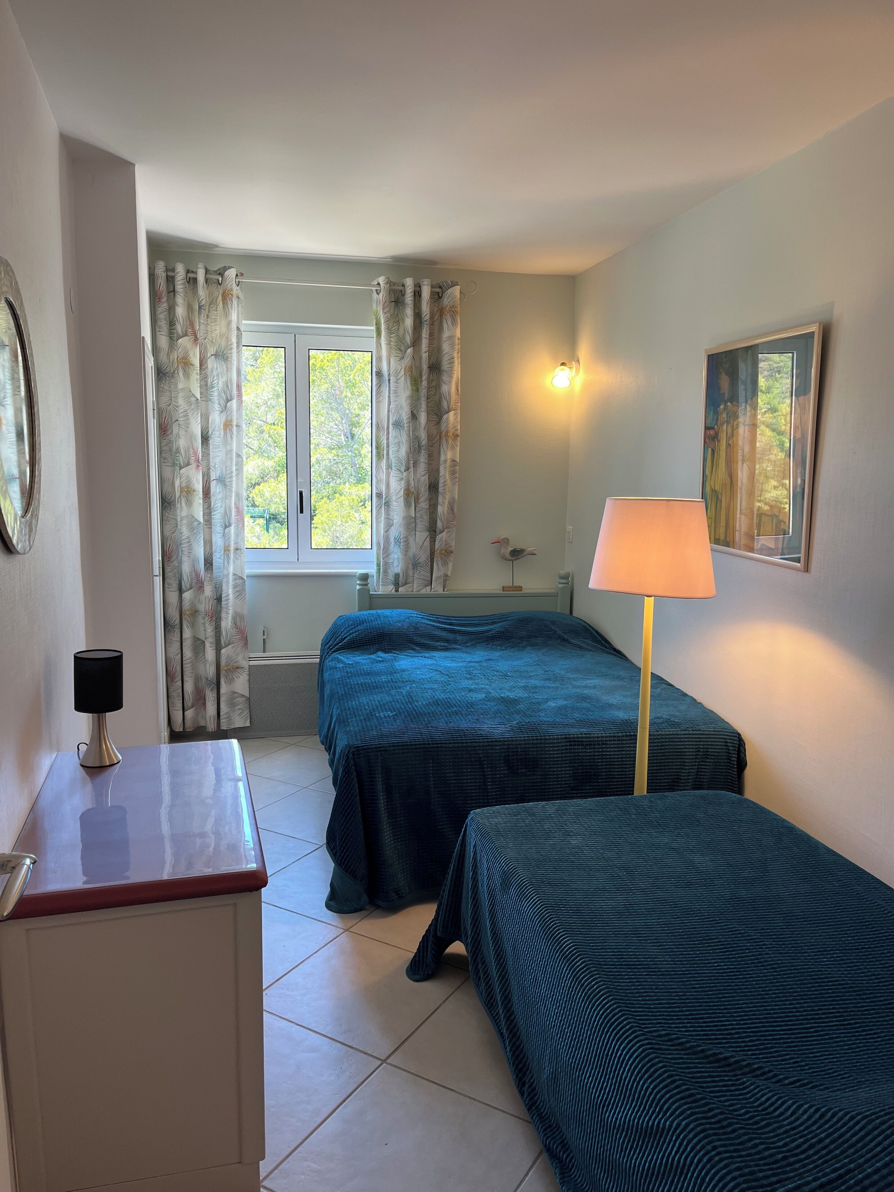 A room with two beds, a desk, a lamp, a painting, and a window with curtains.