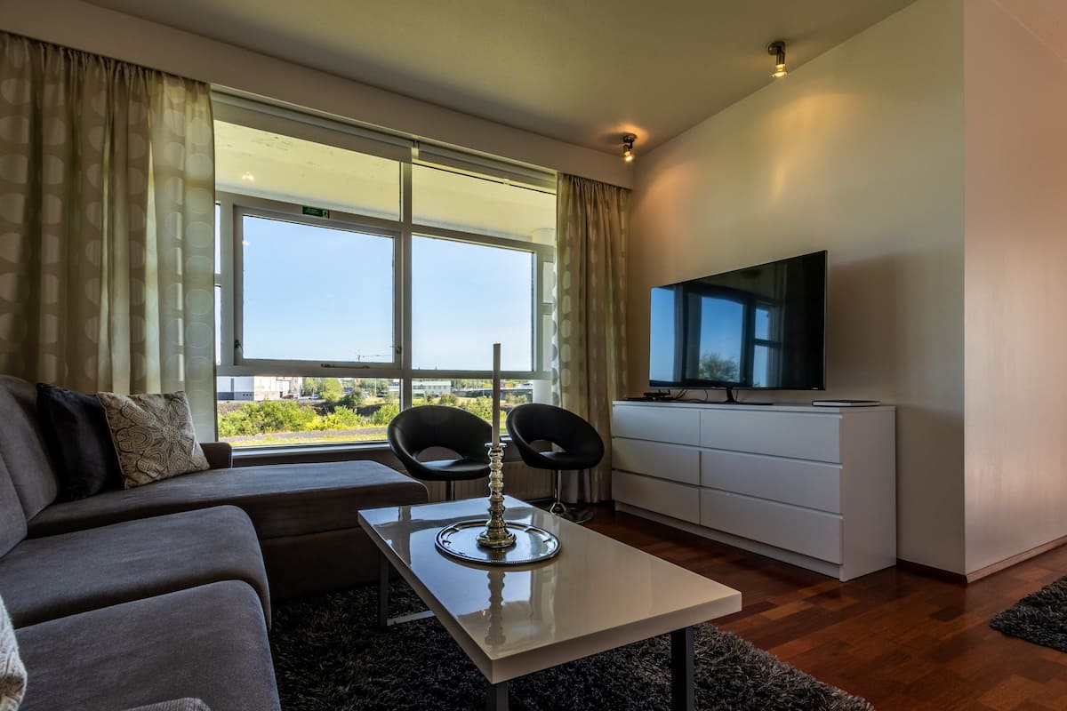 Una sala de estar moderna con un sofá, dos sillones, una mesa de centro, un televisor y un amplio ventanal con cortinas.