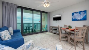 A modern living room with a blue sofa, a dining table with chairs, a flat-screen TV, and a large window overlooking a balcony.