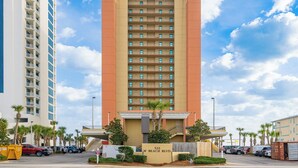 A multi-story hotel with a central entrance, balconies, and a parking area with cars and palm trees.
