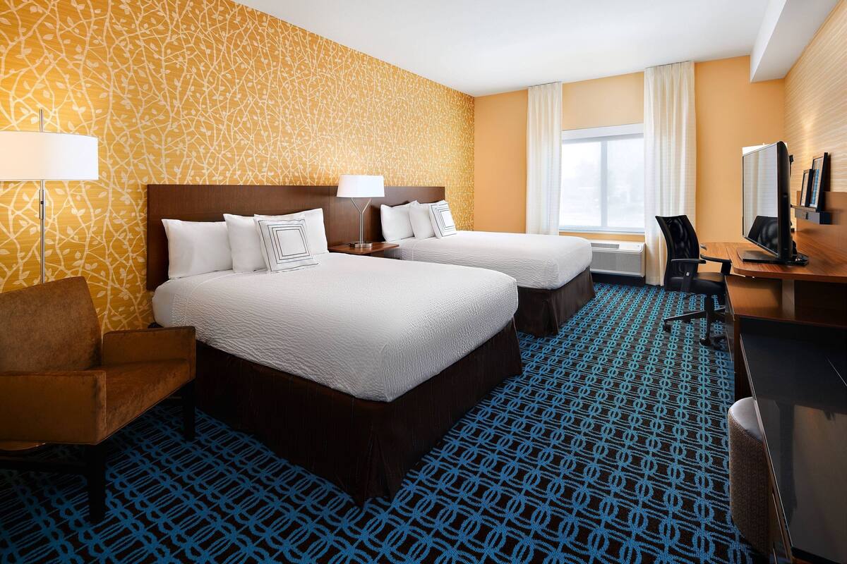Habitación de hotel con dos camas, un escritorio, una silla, una lámpara y un ventanal grande.