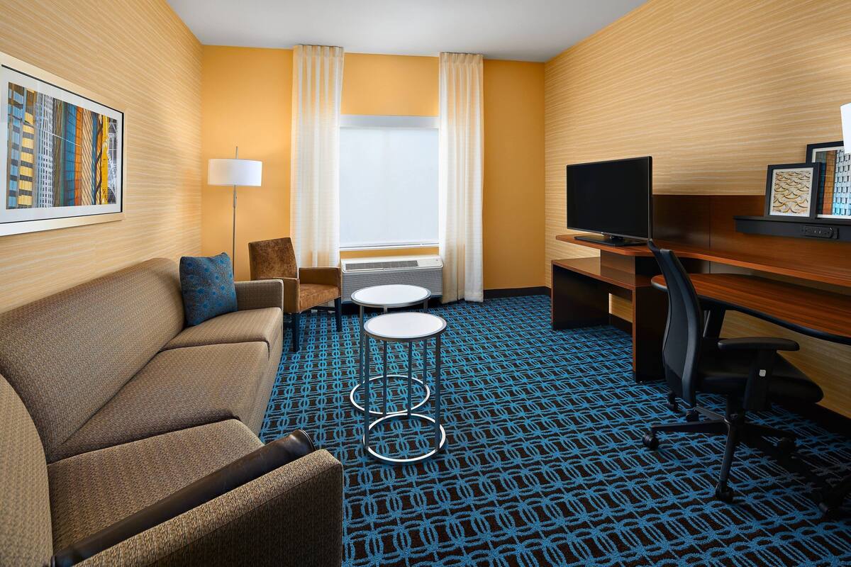 Habitación de hotel con sofá, escritorio con televisor, una mesita pequeña y alfombra estampada.