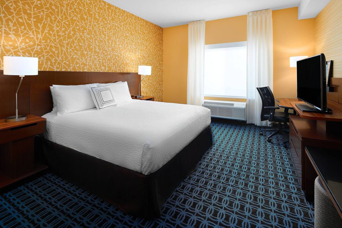 Habitación de hotel con una cama grande, dos mesitas de noche con lámparas, un escritorio con silla, un televisor y una ventana con cortinas.