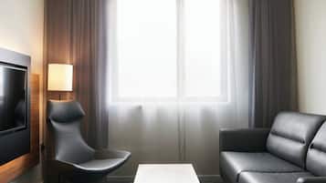 Una camera d'albergo moderna con una TV a schermo piatto, un divano grigio, una sedia dalle linee originali e una grande finestra con tende leggere.