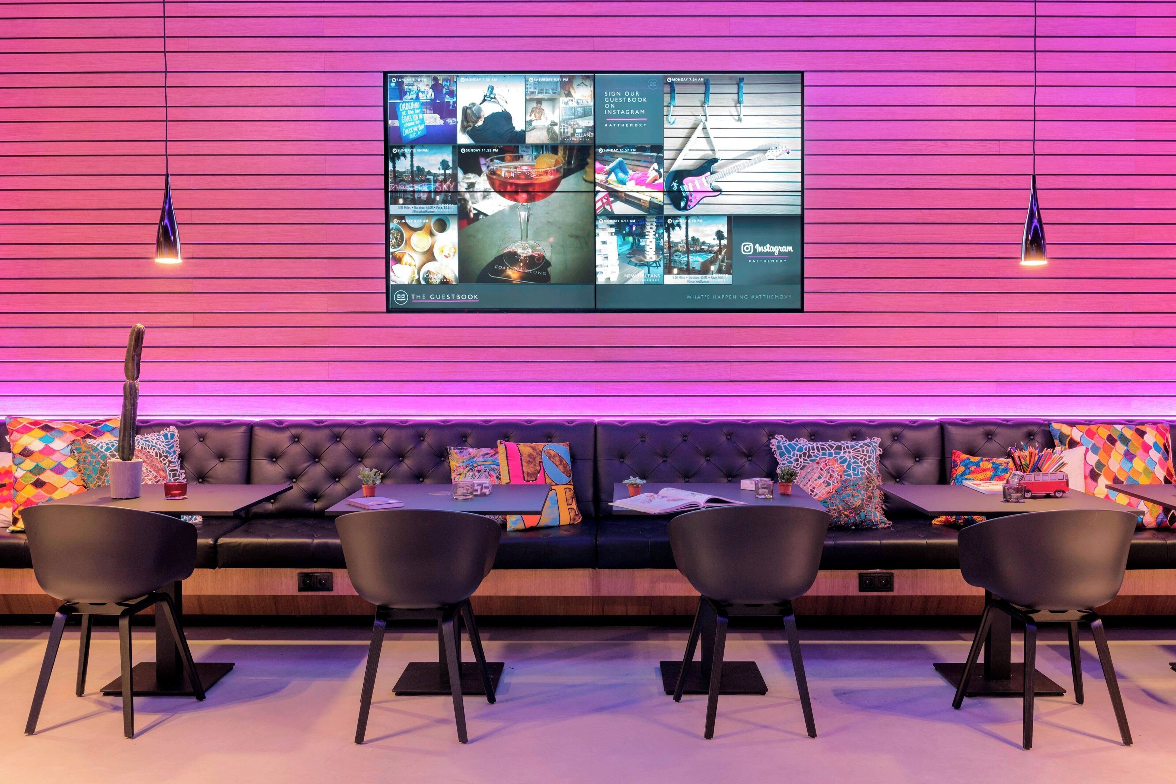 Un área de descanso moderna con un televisor de pantalla plana grande mostrando diverso contenido, un sofá negro y cómodo con cojines de colores, y sillas negras alrededor de una mesita.