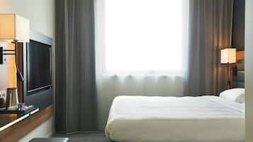 A hotel room with a bed, a TV, a desk with a lamp, and a window with curtains.