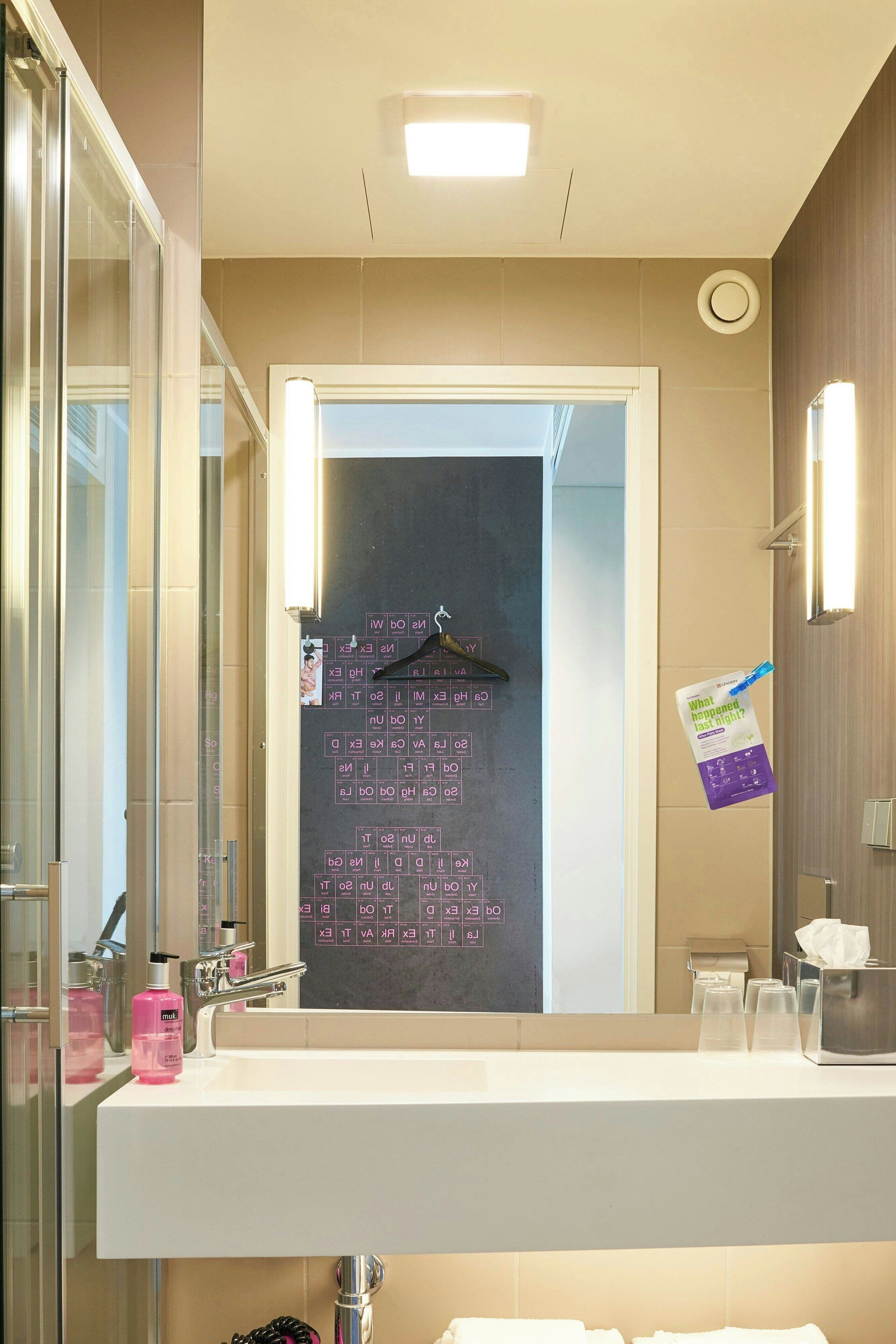Un baño con un espejo grande, un lavabo y una zona de ducha. Hay artículos de aseo en la encimera del lavabo y un perchero encima del espejo.