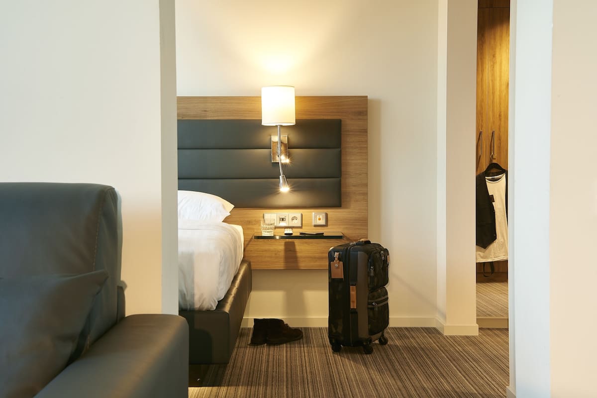 A hotel room with a bed, a bedside table with a lamp, a suitcase, and a coat rack.