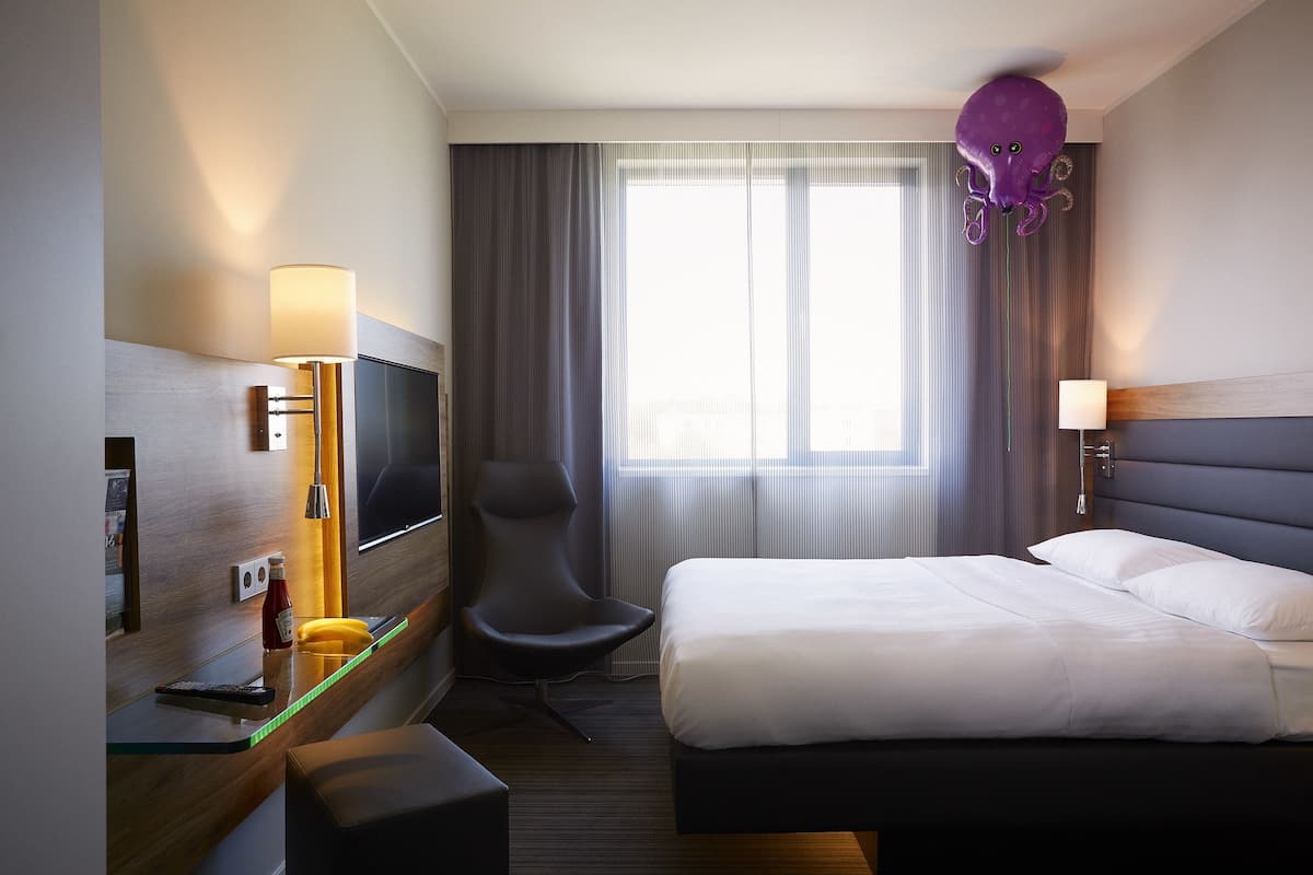 A hotel room with a bed, a desk with a lamp, a chair, a TV, and a window with curtains.