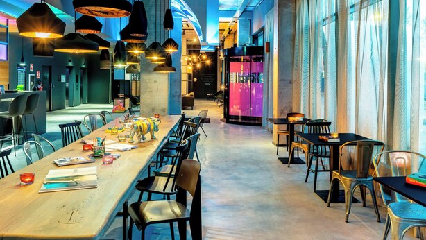 A modern restaurant with a long communal table, multiple hanging pendant lights, and a view of the city through large windows. - Moxy Milan Linate Airport (Segrate)
