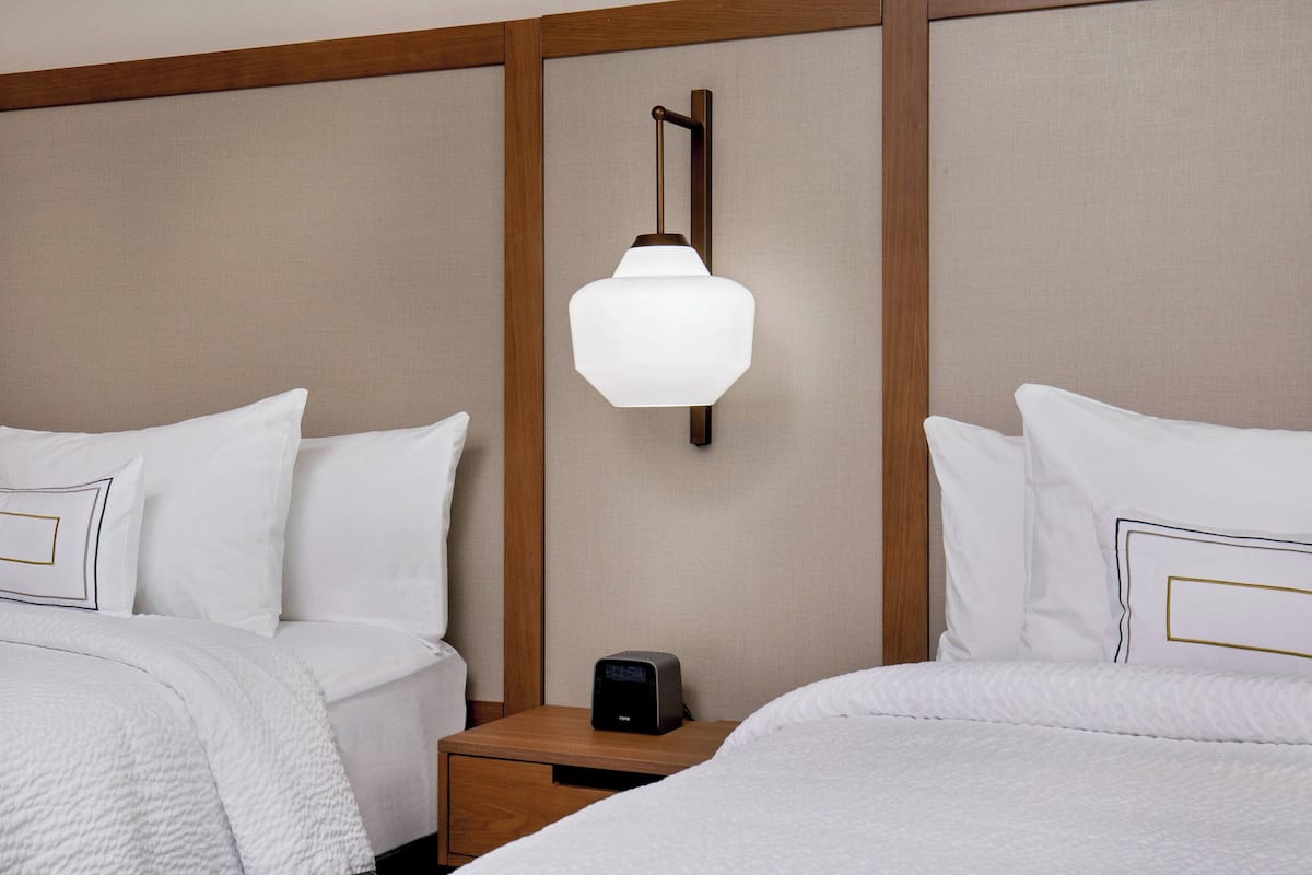 A hotel room with two beds, a nightstand with a lamp, and a digital clock.