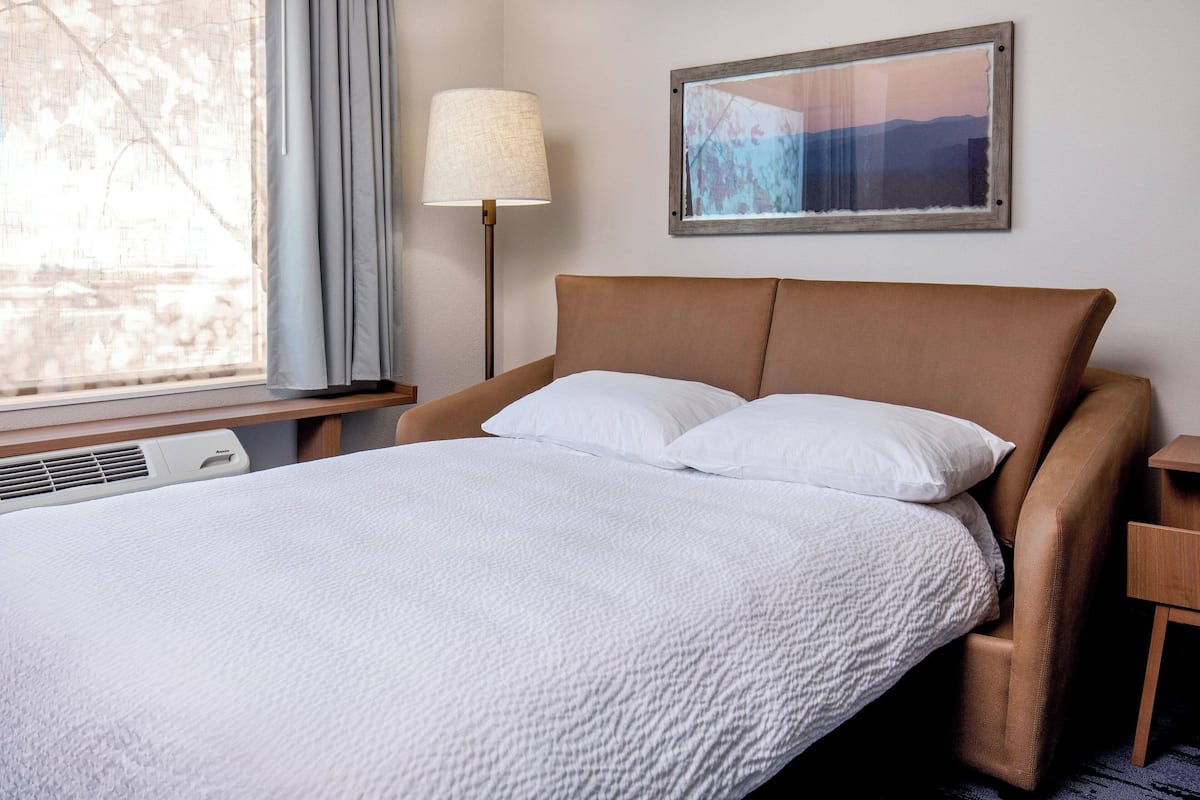 A neatly made bed with white bedding, a brown headboard, a bedside lamp, and a framed picture on the wall.