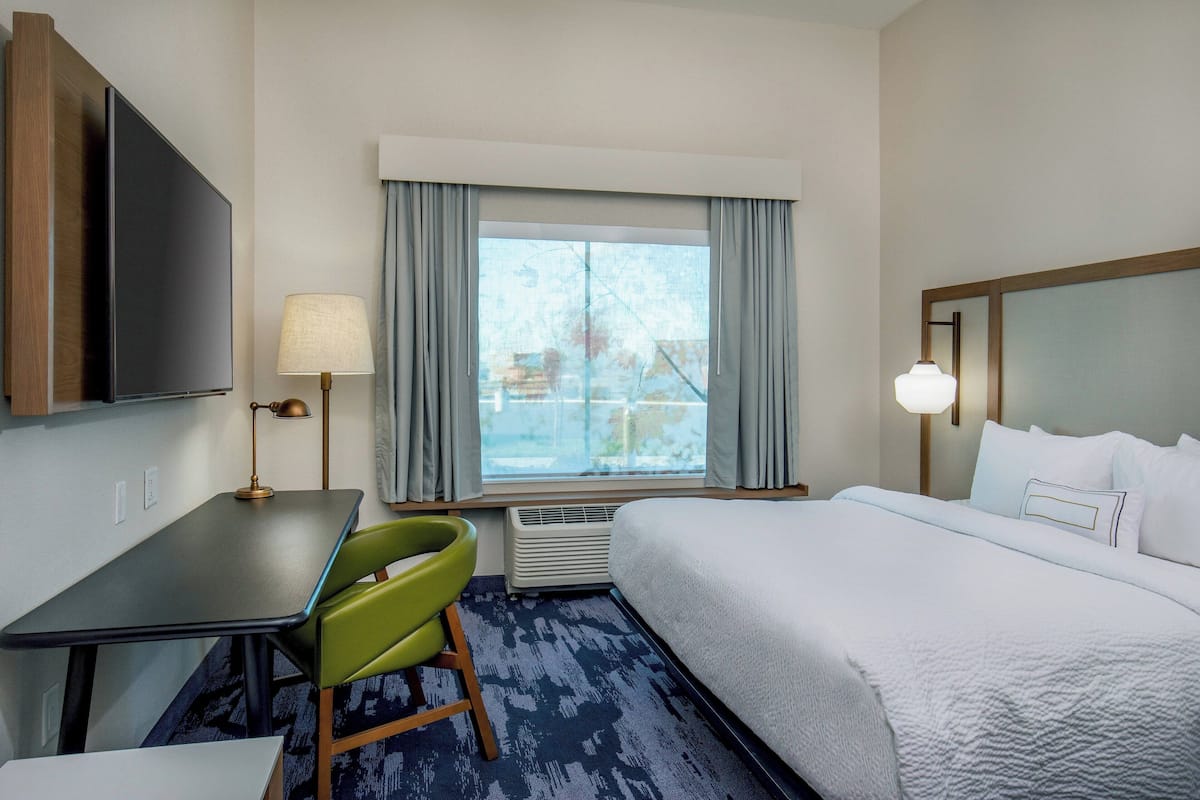 A hotel room with a bed, a desk, a chair, a TV, a window with curtains, and a lamp.
