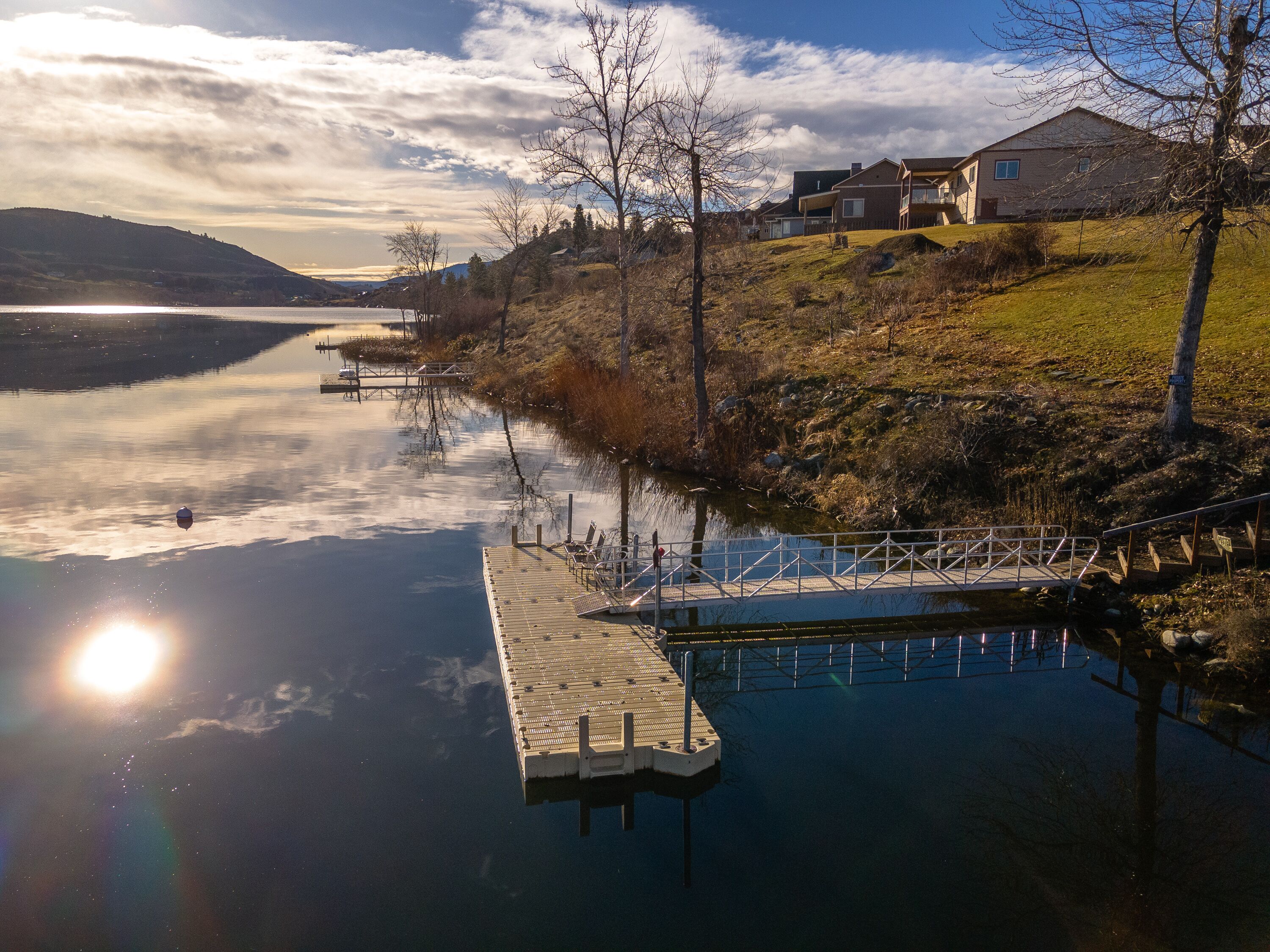 A calm lake with a dock extending into the water, surrounded by hills and a few houses in the background.