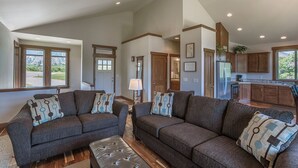 A spacious living room with two sofas and a coffee table, a kitchen with wooden cabinets, and large windows offering a view of the outdoors. - Wapato Lake Retreat-Sleeps 6, Waterfront and dock! (Manson)