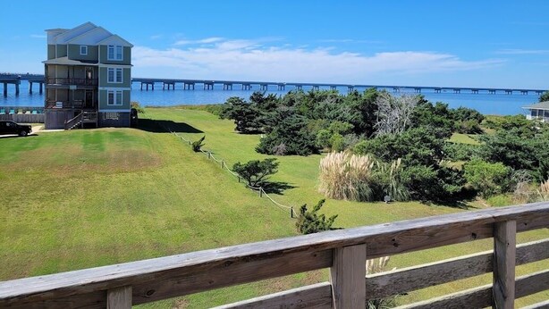 A multi-story building with a balcony, a grassy area, and a wooden fence. - OBX Beach House + Bikes-Paddle Boards-Kayaks-Beach Chairs-Great Fishing! (Rodanthe)