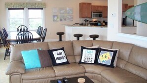 A living room with a beige sectional sofa, a dining table with chairs, a kitchen area with wooden cabinets and a microwave, and a ceiling fan. - OBX Beach House + Bikes-Paddle Boards-Kayaks-Beach Chairs-Great Fishing! (Rodanthe)