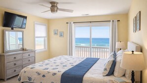 A bedroom with a bed, a dresser, a TV, a ceiling fan, a window with a view of the ocean, and a nightstand with a lamp.